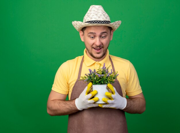 Foto gratuita joven jardinero vistiendo mono y sombrero en guantes de trabajo sosteniendo la planta en maceta mirándola sorprendida de pie sobre la pared verde