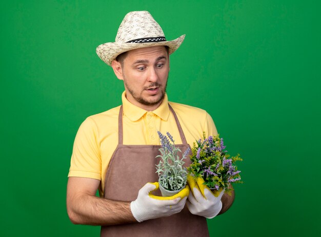 Joven jardinero vistiendo mono y sombrero en guantes de trabajo sosteniendo plantas en macetas mirándolas confundirse de pie sobre la pared verde