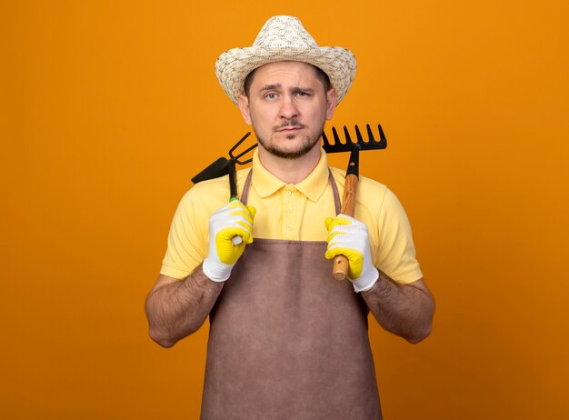 Joven jardinero vistiendo mono y sombrero sosteniendo azadón y mini rastrillo mirando al frente con cara seria de pie sobre la pared naranja