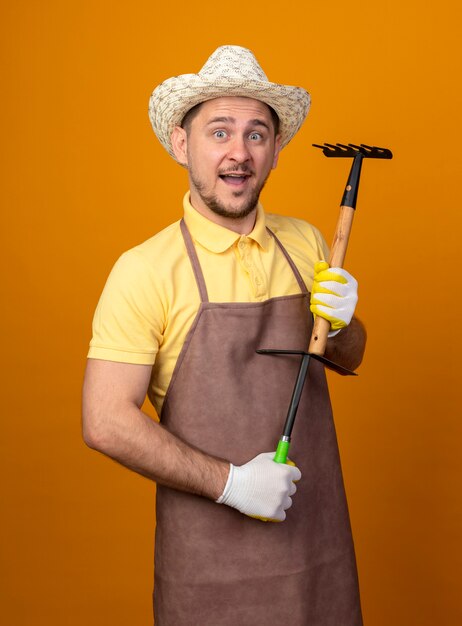 Joven jardinero vistiendo mono y sombrero sosteniendo mini rastrillo mirando al frente feliz y sorprendido de pie sobre la pared naranja