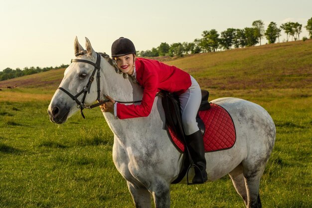 Joven jinete, vestida con redingote rojo y calzones blancos, con su caballo a la luz del atardecer. Fotografía al aire libre en ambiente de estilo de vida