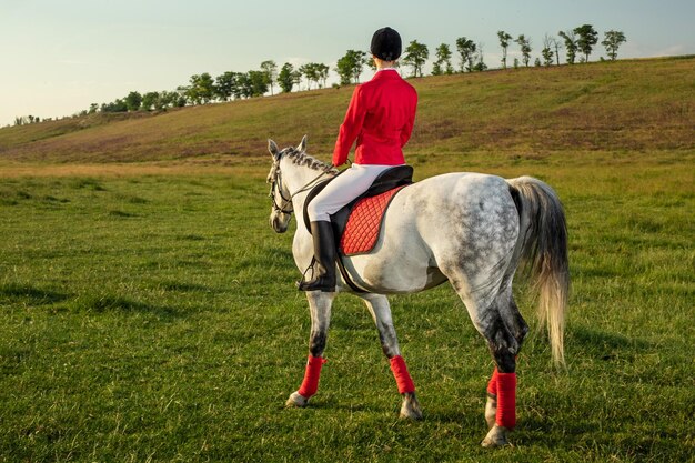 Joven jinete, vestida con redingote rojo y calzones blancos, con su caballo a la luz del atardecer. Fotografía al aire libre en ambiente de estilo de vida