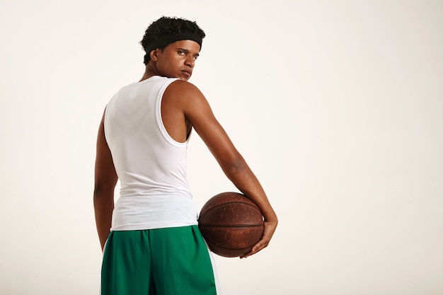 Joven jugador de baloncesto afroamericano serio decidido en uniforme blanco y verde con un afro corto sosteniendo una vieja pelota de baloncesto marrón en la cadera y mirando hacia atrás
