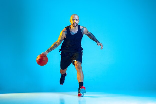 Joven jugador de baloncesto del equipo vistiendo ropa deportiva, practicando en acción, aislado en la pared azul en luz de neón