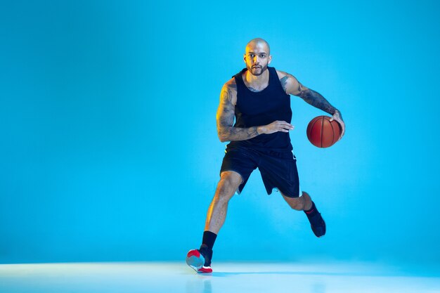 Joven jugador de baloncesto del equipo vistiendo ropa deportiva, practicando en acción, movimiento aislado sobre fondo azul en luz de neón. Concepto de deporte, movimiento, energía y estilo de vida dinámico y saludable.