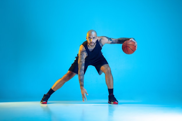 Joven jugador de baloncesto del equipo vistiendo ropa deportiva, practicando en acción, movimiento aislado sobre fondo azul en luz de neón. Concepto de deporte, movimiento, energía y estilo de vida dinámico y saludable.