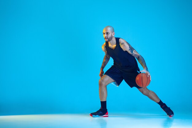 Joven jugador de baloncesto del equipo vistiendo ropa deportiva, practicando en acción, movimiento aislado sobre fondo azul en luz de neón. Concepto de deporte, movimiento, energía y estilo de vida dinámico y saludable.