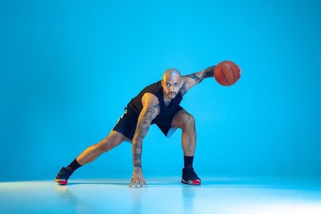 Joven jugador de baloncesto del equipo vistiendo ropa deportiva, practicando en acción, movimiento aislado sobre fondo azul en luz de neón. Concepto de deporte, movimiento, energía y estilo de vida dinámico y saludable.