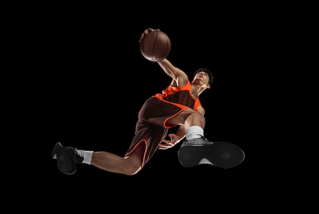 Joven jugador de baloncesto profesional en acción, movimiento aislado en la pared negra, mira desde abajo. Concepto de deporte, movimiento, energía y estilo de vida dinámico y saludable. Entrenando, practicando.