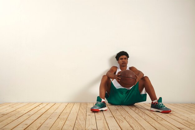 Joven jugador de baloncesto serio en pantalones cortos verdes y camisa blanca tomando un descanso contra una pared blanca sosteniendo una pelota de baloncesto grunge en sus manos
