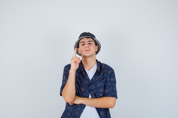 Joven levantando el dedo índice en gesto de eureka mientras sostiene la mano debajo del codo en camiseta blanca, camisa floral, gorra y luciendo sensato. vista frontal.