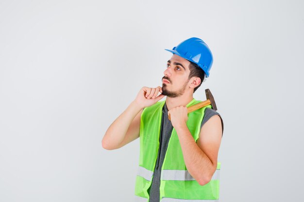 Joven levantando el hacha por encima del hombro y la barbilla inclinada en la mano en uniforme de construcción y mirando pensativo