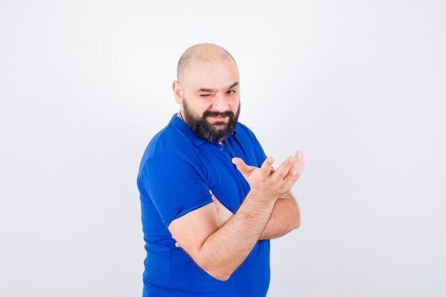 Joven levantando la mano con la palma abierta a un lado mientras guiña un ojo con la camisa azul, vista frontal.