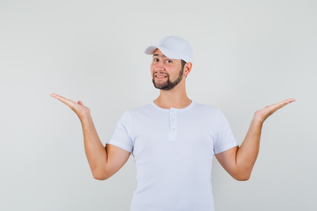 Joven levantando las manos como sosteniendo o mostrando algo en camiseta blanca, gorra y mirando alegre, vista frontal.