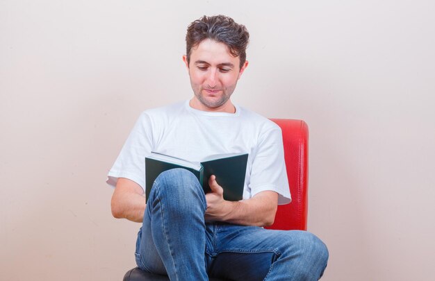 Joven leyendo el libro mientras está sentado en una silla en camiseta, jeans y mirando alegre