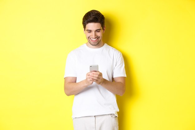 Joven leyendo un mensaje de texto en el teléfono inteligente, mirando la pantalla del teléfono móvil y sonriendo, de pie con una camiseta blanca sobre fondo amarillo