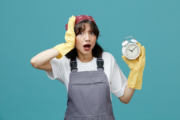 Una joven limpiadora ansiosa que usa pañuelo uniforme y guantes de goma que muestran el despertador con la mano en la cabeza mirando la cámara aislada en el fondo azul