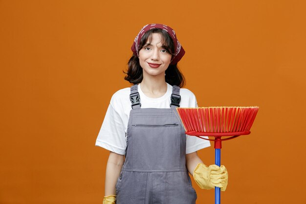 Una joven limpiadora complacida con guantes de goma uniformes y un pañuelo sosteniendo un trapeador mirando a la cámara aislada en un fondo naranja