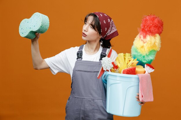 Una joven limpiadora concentrada con uniforme y pañuelo sosteniendo una esponja en el aire y un cubo de herramientas de limpieza mirando a un lado simulando limpiar algo con una esponja aislada en un fondo naranja
