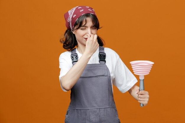 Una joven limpiadora disgustada con uniforme y pañuelo sosteniendo y mirando el émbolo haciendo un gesto de mal olor aislado en el fondo naranja