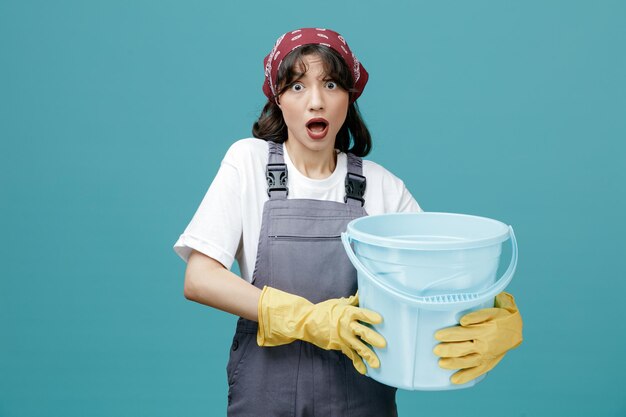 Una joven limpiadora impresionada con un pañuelo uniforme y guantes de goma sosteniendo un balde con ambas manos mirando la cámara aislada de fondo azul
