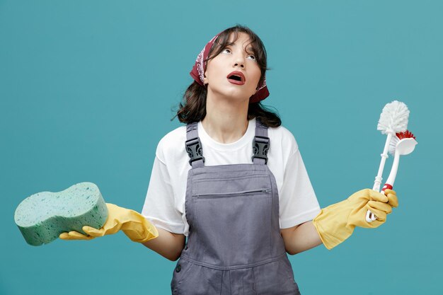 Una joven limpiadora impresionada con un pañuelo uniforme y guantes de goma sosteniendo una esponja y cepillos de baño mirando hacia arriba aislada en el fondo azul