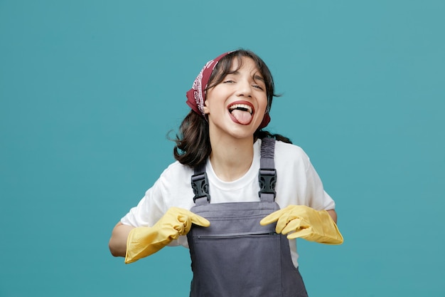 Joven limpiadora juguetona con pañuelo uniforme y guantes de goma agarrando su uniforme mirando a la cámara mostrando la lengua aislada en el fondo azul