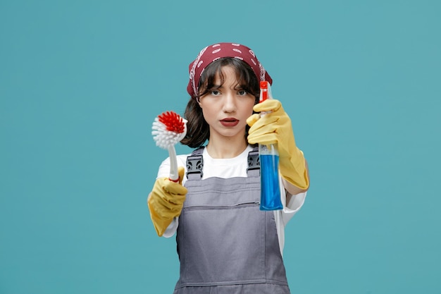 Una joven limpiadora seria con pañuelo uniforme y guantes de goma mirando la cámara estirando el cepillo y el limpiador hacia la cámara aislada en el fondo azul