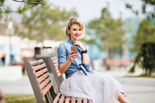 Joven linda chica rubia con pelo corto sentada en un banco de madera sosteniendo un teléfono inteligente y sonriendo con camisa azul denim.