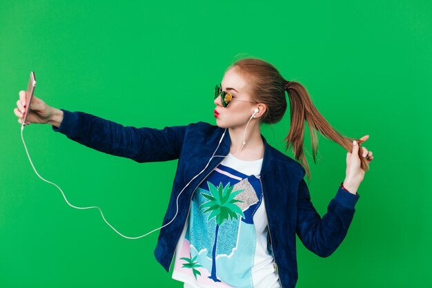 Una joven linda con cola de pelo largo está haciendo un selfie cerca de la pared verde en el fondo. Lleva gafas de sol con corazones y tiene labios rojos. Ella está escuchando música en auriculares.