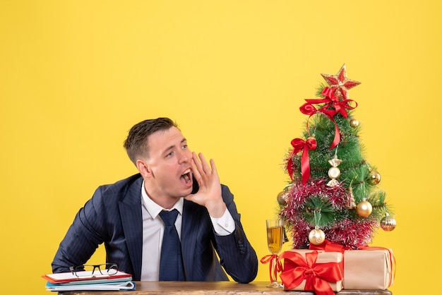 joven llamando a alguien sentado en la mesa cerca del árbol de navidad y regalos en amarillo