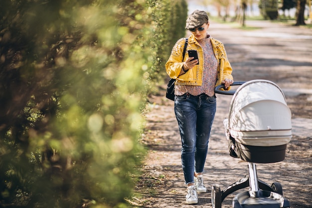 Foto gratuita joven madre caminando con carro de bebé en el parque y usando el teléfono