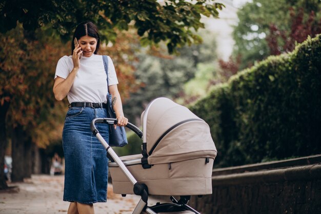 Joven madre caminando con cochecito en el parque y hablando por teléfono