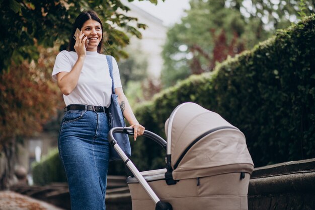 Joven madre caminando con cochecito en el parque y hablando por teléfono