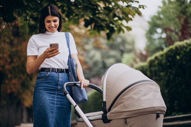 Joven madre caminando con cochecito en el parque y hablando por teléfono