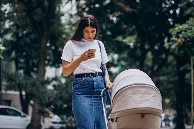 Joven madre caminando en el parque con cochecito de bebé y hablando por teléfono