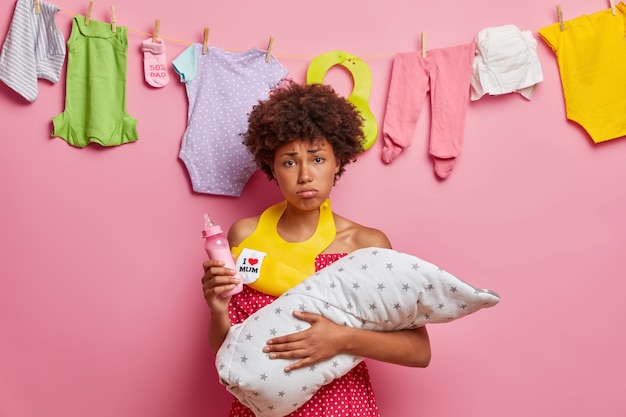 Joven madre cansada disgustada sostiene al bebé envuelto en una manta sostiene el biberón usa babero alrededor del cuello va a alimentar al recién nacido tiene mucho trabajo en la casa. Mamá amamantando a su hija pequeña. Familia monoparental