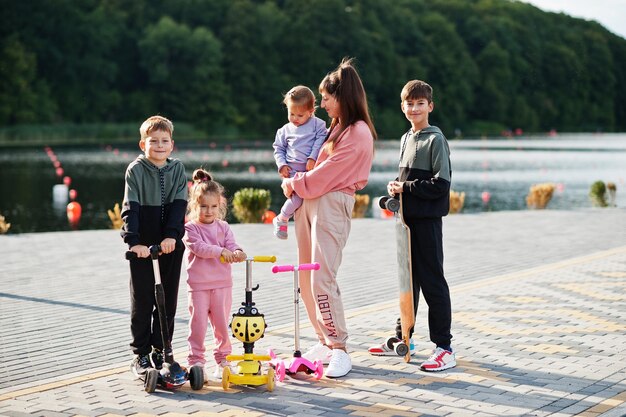 Joven madre elegante con cuatro hijos al aire libre La familia deportiva pasa tiempo libre al aire libre con scooters y patines