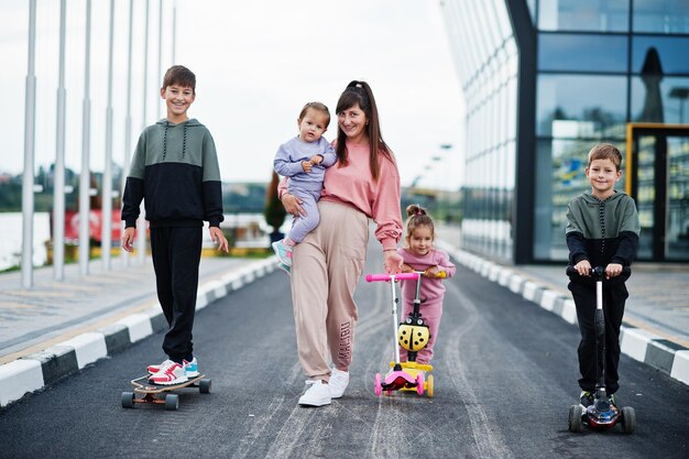 Joven madre elegante con cuatro hijos al aire libre La familia deportiva pasa tiempo libre al aire libre con scooters y patines