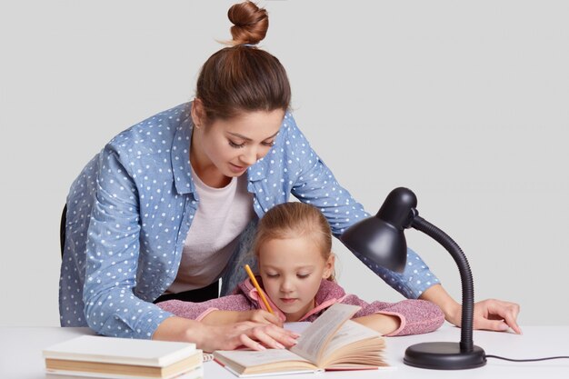 La joven madre con experiencia se inclina cerca de su hijo pequeño, ayuda a hacer la tarea del hogar, muestra qué reescribir en el libro, rodeada de una lámpara de lectura, aislada en blanco