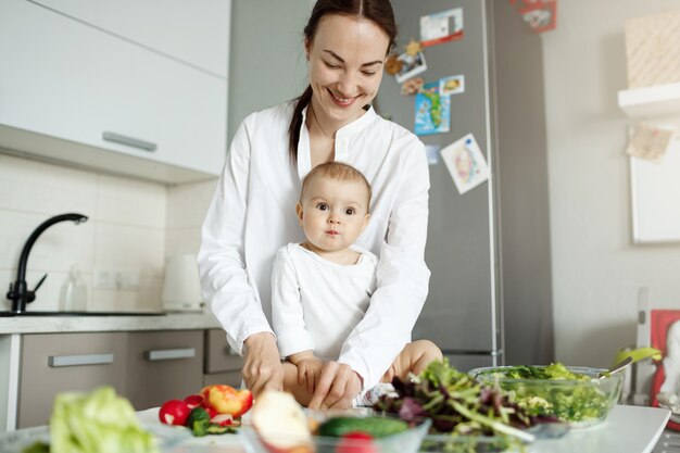 Joven madre feliz cocinando comida en la cocina y cuidando al bebé