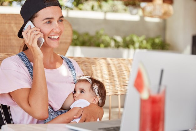 Joven madre feliz con gorra elegante y ropa informal, amamanta a su pequeño hijo, le da leche materna, habla con alguien a través de un teléfono inteligente y mira videos para padres sin experiencia en una computadora portátil