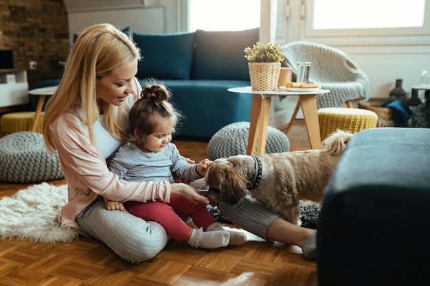 Joven madre feliz y su pequeña hija divirtiéndose con su perro en la sala de estar