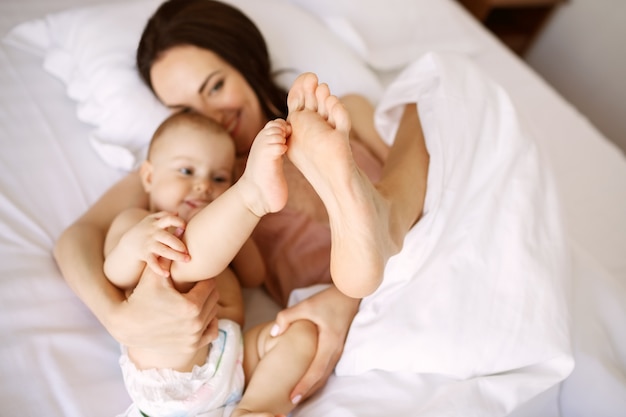 Joven madre hermosa y bebé recién nacido acostado en la cama sonriendo engañando en casa. Desde arriba.