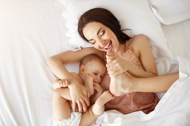 Joven madre hermosa y bebé recién nacido acostado en la cama sonriendo engañando en casa. Desde arriba.