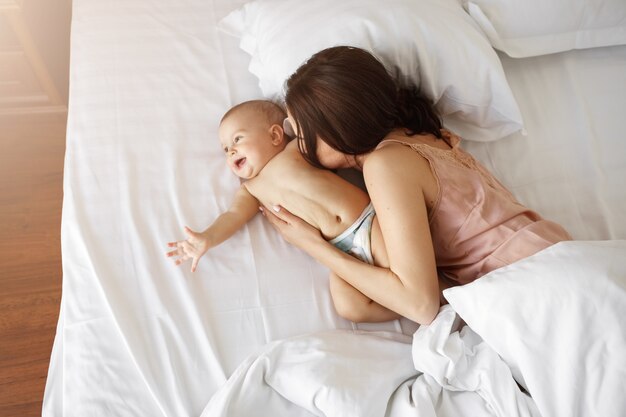 Joven madre hermosa y bebé recién nacido acostado en la cama sonriendo engañando en casa. Desde arriba.