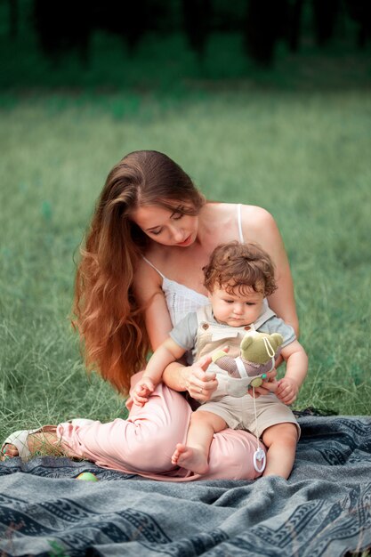 Joven madre hermosa sentada con su pequeño hijo contra la hierba verde. Mujer feliz con su bebé en un día soleado de verano. Familia caminando por el prado.
