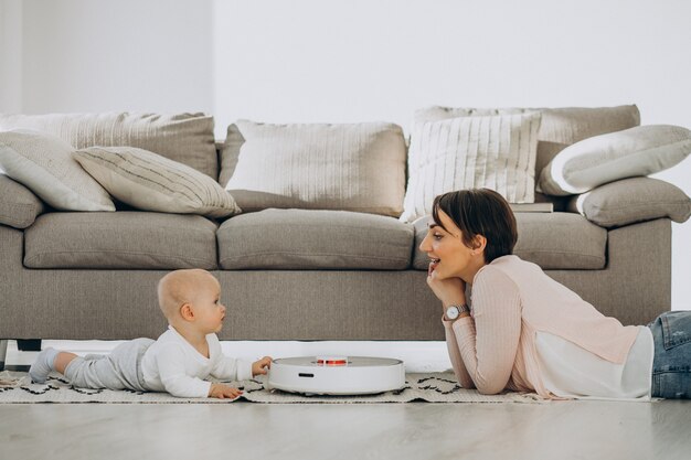 Joven madre con un hijo pequeño y viendo robot aspirador haciendo tareas domésticas