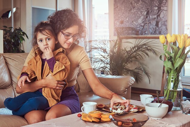 Joven madre con una linda hijita desayunando en el sofá de casa.