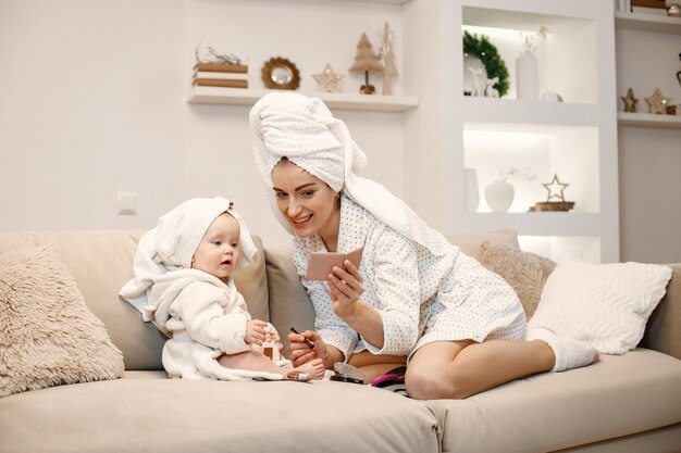 Joven madre y niña pequeña vistiendo batas con cabello envuelto en toallas Mujer y niña sentadas en un sofá Mujer sosteniendo cosméticos decorativos